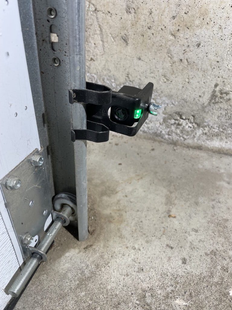 Garage door opener safety sensor showing a green light to indicate proper sensor operation in Vaughan by Penguin Garage Doors.